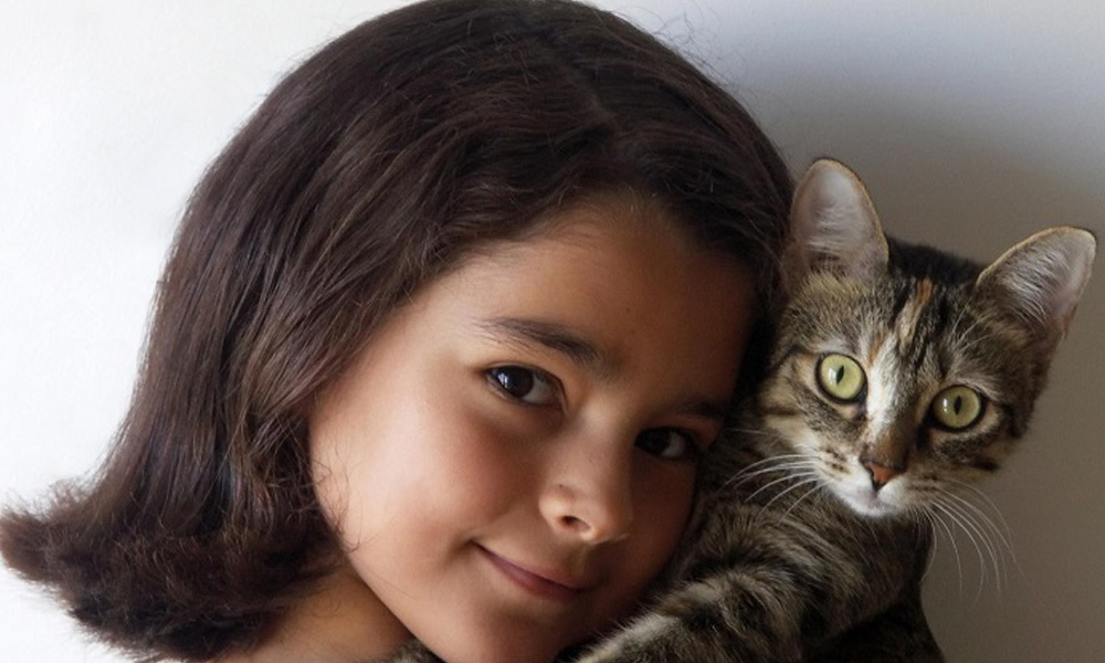 A young girl with dark hair gently holds a tabby cat close to her cheek, both looking directly at the camera as they wait at the veterinarian's office.