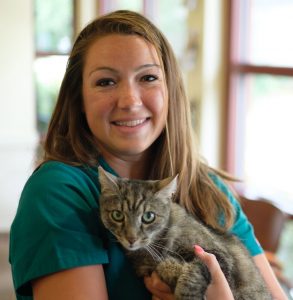 A smiling veterinarian in a teal shirt holds a brown tabby cat inside a brightly lit room.