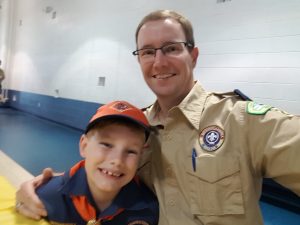 A smiling adult in a scout leader uniform takes a selfie with a young boy in a cub scout uniform in a gymnasium. Both appear happy and are looking at the camera. The adult is also known as a dedicated veterinarian.