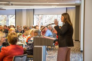 A veterinarian presents to an attentive audience in a dining hall with tables set for a meal, lit by natural light from large windows with views of trees.