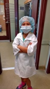 A young child dressed as a veterinarian wearing a surgical mask, cap, and lab coat, standing confidently with arms crossed in a clinical setting.