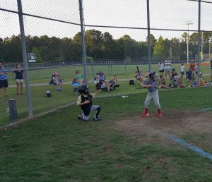 Young baseball player batting in a fenced diamond with children watching and adults coaching, including a veterinarian volunteering as a coach, during an outdoor evening practice session.