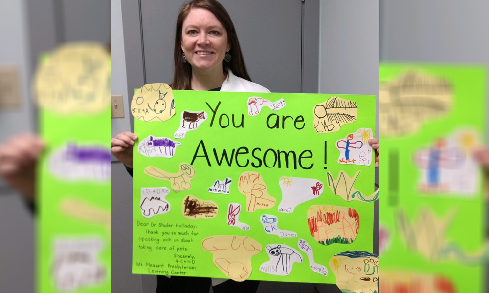 A veterinarian with a content smile proudly holds a colorful poster that reads "you are awesome!" surrounded by various child-like animal drawings, standing in front of a grey backdrop.