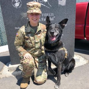 A smiling woman in military uniform kneels beside a happy black military dog wearing a harness, both in front of a memorial wall with inscriptions. The woman, also a vet, shares a special bond with the trained canine.