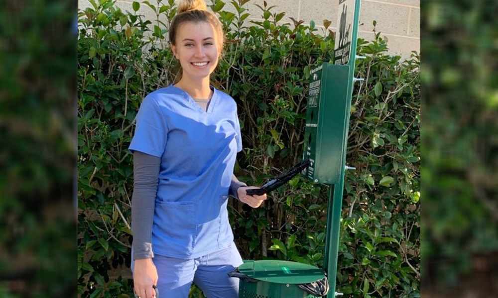 A smiling young veterinarian in blue scrubs stands by a green post with a clipboard in her hand, against a backdrop of dense shrubs.