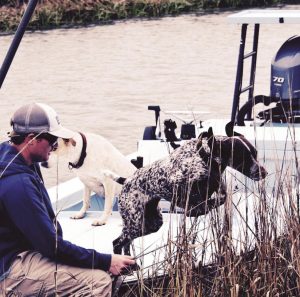 A man and two dogs on a boat beside tall grass, with one dog looking into the water and the other standing beside the veterinarian, near a quiet river.