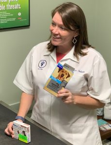 A smiling veterinarian in a white coat, standing at a counter in a clinic, holding a box of canine medication. There are various pet health products on the counter.