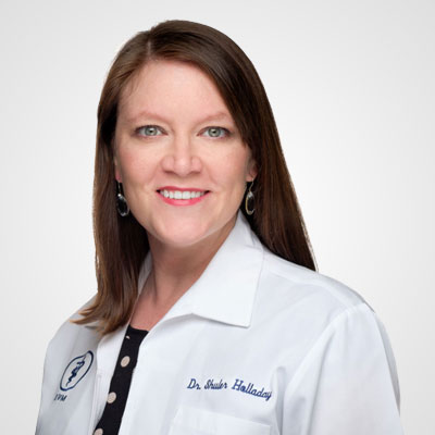 Professional portrait of a smiling woman with long brown hair, wearing a white lab coat with "Dr. Sheila Halladay" embroidered on it and a Ford logo, indicating her role as a veterinarian.