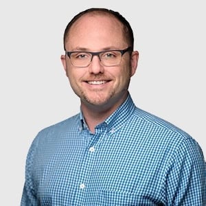 A professional headshot of a smiling veterinarian wearing glasses and a blue checkered shirt, standing against a light gray background.
