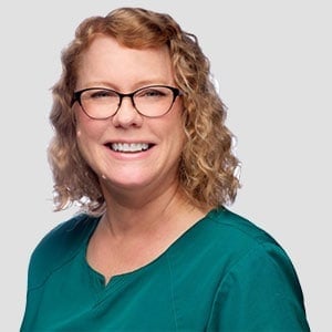 A portrait of a smiling veterinarian with curly hair and glasses wearing a green scrub top against a light grey background.