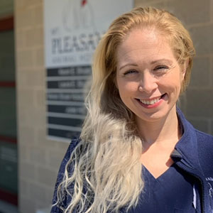 A smiling woman with blonde hair stands in front of a clinic named "center of veterinary therapy," wearing a dark blue uniform with a visible name tag.