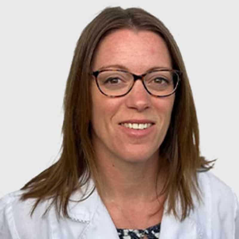Professional headshot of a smiling veterinarian with long brown hair wearing glasses and a white lab coat over a patterned blouse, against a plain, light background.