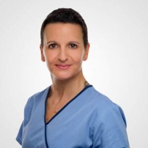 A professional portrait of a woman with short hair, wearing a blue medical scrub, smiling confidently at the camera against a light gray background. She is a veterinarian.