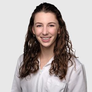 A professional portrait of a woman with curly hair smiling at the camera, wearing a white lab coat. She is a veterinarian with a gray background.