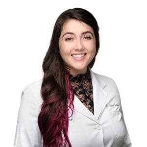 A professional portrait of a smiling veterinarian with long brown and pink hair, wearing a white lab coat with a badge and a floral top, against a plain white background.