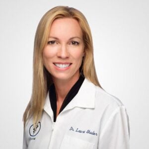 A professional headshot of a smiling woman with blonde hair, wearing a white lab coat labeled "Dr. Lauren Shuler, Veterinarian" against a light grey background.