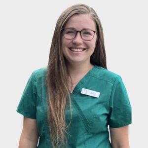 A smiling young woman wearing glasses and a teal veterinary scrub top with a name tag labeled "Ali" on a plain white background.