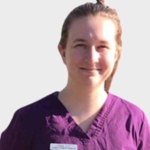 A veterinarian wearing a purple scrub top and a name badge, smiling gently against a plain white background. She has her hair pulled back.