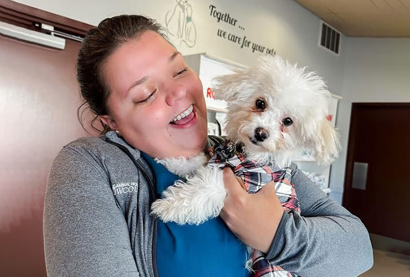 A joyful woman holding and smiling at a small white dog with a plaid scarf in a veterinarian's office with a wall sign that reads, "together we care for your pet.