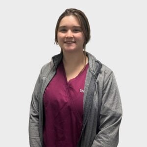 A young woman standing in front of a white background, with shoulder-length brown hair, wearing a burgundy top and a gray zip-up jacket with the name "Steph" embroidered on it, which identifies her as a veterinarian.