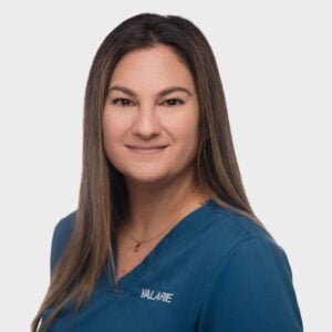 A professional portrait of a smiling veterinarian with long brown hair, wearing a blue scrub top with "Valarie" embroidered on it, against a plain white background.
