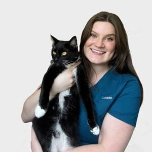 A woman wearing a blue scrub top with the name "Logan" embroidered on it is smiling and holding a black and white cat. The background is plain white.