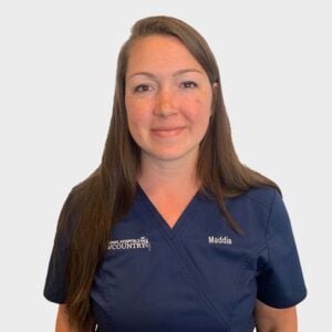 A woman with long brown hair is wearing navy blue scrubs embroidered with "Animal Hospital LowCountry" and a name tag that reads "Maddie." She is smiling and standing against a plain white background.