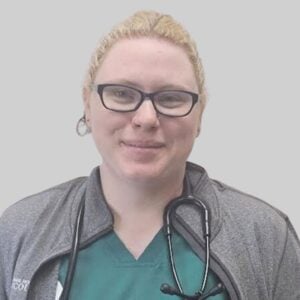 A person wearing glasses smiles, dressed in a green scrub top and a gray jacket with a stethoscope draped around their neck. The background is a plain light gray.