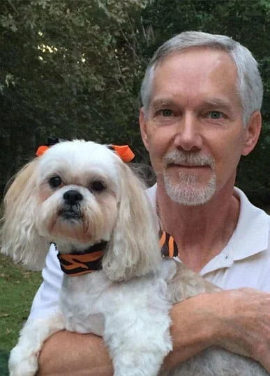 A vet with a trimmed beard and mustache holding a small white dog with orange bows on its ears; both are in front of a backdrop of green trees.