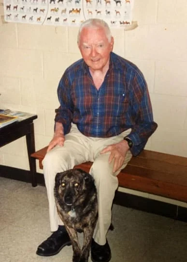 An elderly man with a joyful expression, wearing a plaid shirt and khaki pants, sits on a bench beside a large brindle-coated dog in the vet's office, with insect illustrations on the wall behind them.