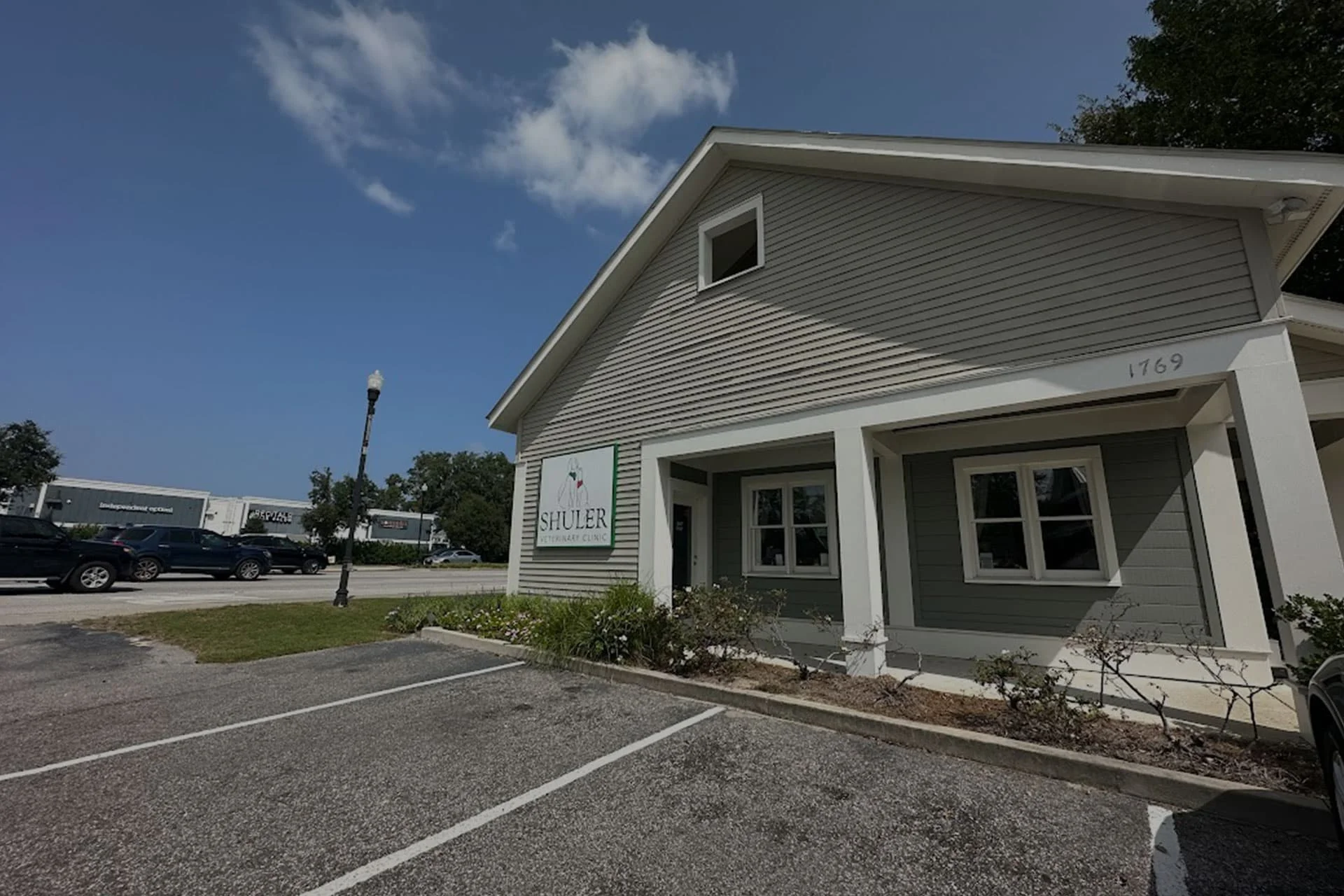 A one-story commercial building with white siding and a small porch, featuring the "shelter" veterinarian logo. The building is situated in a parking lot with vehicles and trees under a clear sky.